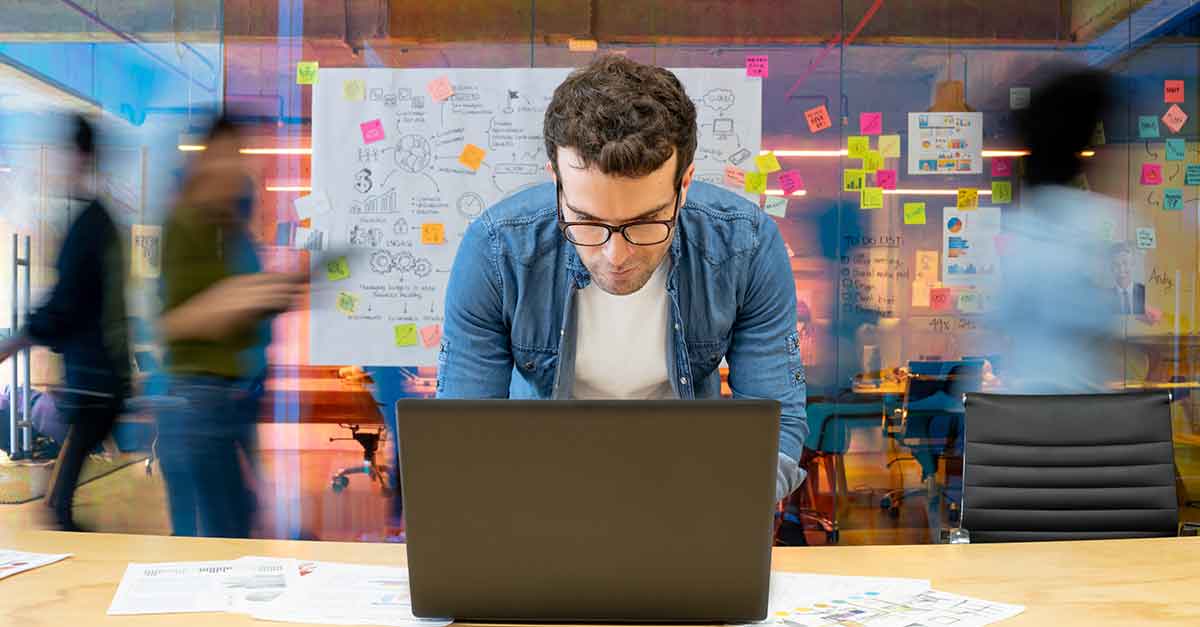 Man working at a creative office using his computer and people moving in the background